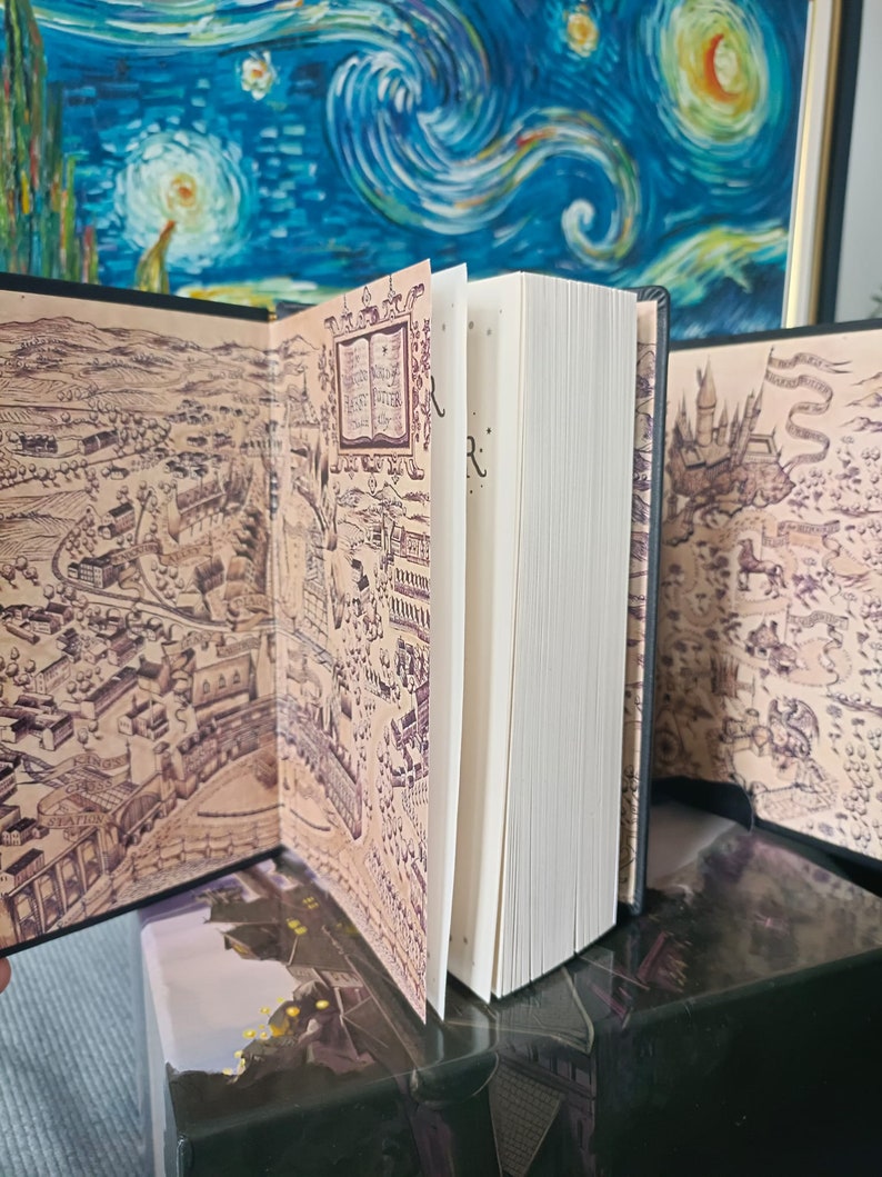 May include: An open book with detailed map illustrations on the inside covers. The pages are white, and the book is on a dark surface. The map is in brown and beige tones, featuring intricate drawings.