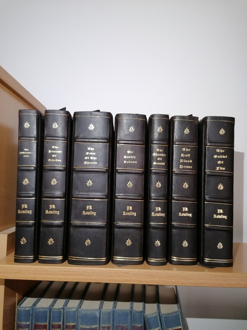May include: A set of seven hardback books with dark brown leather covers and gold lettering. The spines show titles such as "The Prisoner of Azkaban" and "The Goblet of Fire" by J.K. Rowling. The books are displayed on a wooden shelf.