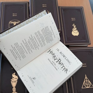 May include: A selection of dark brown hardcover books with gold detailing. One book is open, revealing the title "Harry Potter and the Philosopher's Stone" by J.K. Rowling. The covers are embossed with gold designs and the title "Harry Potter".