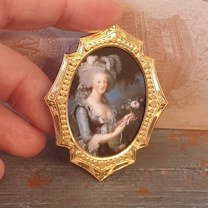 以下が含まれることがあります： 白いドレスと白い羽根の帽子をつけた女性の肖像画が描かれた、金色の楕円形のロケット。ロケットはビーズの縁取りがされており、茶色の木製の表面に置かれている。