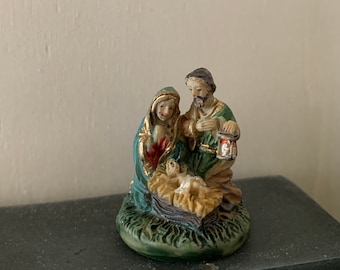 May include: A small, green, and gold figurine of the Holy Family. Mary, Joseph, and baby Jesus are depicted in a nativity scene. The figurine is made of resin and has a detailed finish.