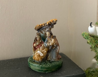 May include: A small, colorful figurine depicting the nativity scene. The figures are made of painted resin and feature Mary, Joseph, and baby Jesus in a manger. The figurine is set against a white background.