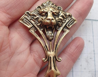 May include: A gold-colored metal brooch with a roaring lion's head at the top. The brooch has an Art Deco design with stylized geometric shapes and a decorative, symmetrical form. The lion's mouth is open, revealing teeth.