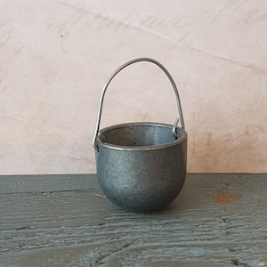 May include: A small, silver-colored metal bucket with a wire handle. The bucket is sitting on a wooden surface.