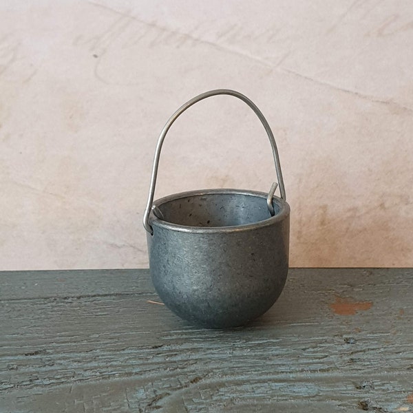 May include: A small, silver-colored metal bucket with a wire handle. The bucket is sitting on a wooden surface.