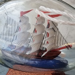 May include: A miniature ship with white and black striped sails is encased in a clear glass bottle. The ship is sitting on a blue base and a wooden stand.