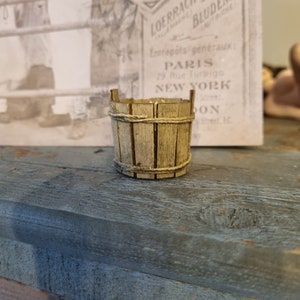 Könnte beinhalten: Ein kleiner Holz-Eimer mit einem Seilgriff. Der Eimer ist aus hellem braunem Holz und hat eine natürliche Oberfläche. Der Seilgriff ist um die Oberseite des Eimers gewickelt.