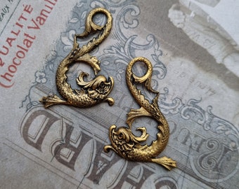 May include: Two antique brass-colored decorative fish ornaments. Each fish has a detailed design with scales, fins, and a curled tail. The ornaments are placed on a vintage paper background with faded text.