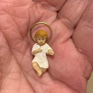 Könnte beinhalten: Kleine Figur des Jesuskindes mit goldenem Heiligenschein. Die Figur trägt ein weißes Gewand und hat hellbraunes Haar. Die Figur ist wahrscheinlich aus Porzellan oder einem ähnlichen Material.