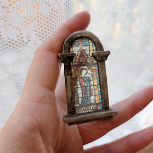 Könnte beinhalten: Ein kleines, steinartiges Miniatur-Bleiglasfenster mit einer religiösen Figur in der Mitte. Das Fenster ist von einem Steinbogen gerahmt und hat ein detailliertes Design.
