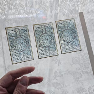 Op de afbeelding: Drie rechthoekige glas-in-loodpanelen met een bloemmotief en een centraal cirkelvormig motief. De panelen hebben een lichtblauw en helder kleurenschema met bruine randen. De panelen worden voor een wit kanten gordijn gehouden.