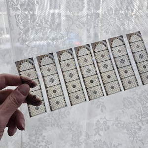 Op de afbeelding: Een hand die een strook kleine, rechthoekige stickers vasthoudt. Elke sticker bevat een gedetailleerde illustratie van een glas-in-loodraam met gewelfde toppen, ingewikkelde patronen en een mix van kleuren. De achtergrond is een wit kanten gordijn.