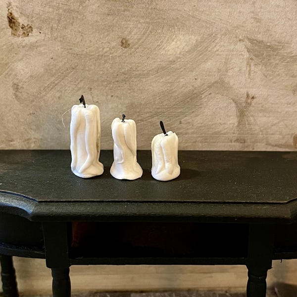May include: Three white candles with black wicks on a black table. The candles are different sizes and have a drippy, melted look.