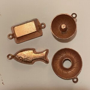 Puede incluir: Cuatro peque&ntilde;os dijes de metal color cobre. Los dijes incluyen una forma rectangular, un pez, un cuenco peque&ntilde;o y una forma de rosquilla. Cada dije tiene un peque&ntilde;o lazo para sujetar.