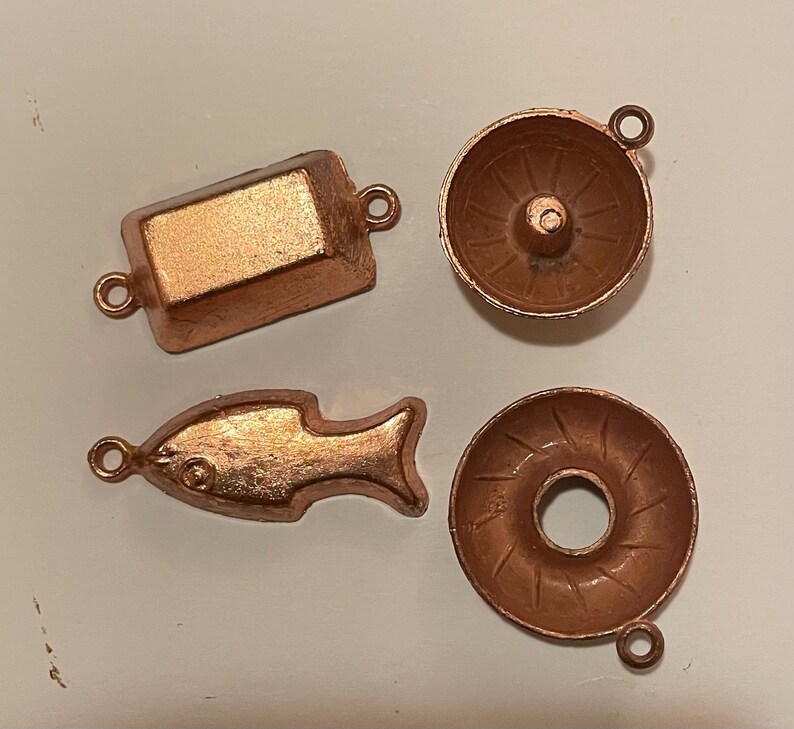 Puede incluir: Cuatro peque&ntilde;os dijes de metal color cobre. Los dijes incluyen una forma rectangular, un pez, un cuenco peque&ntilde;o y una forma de rosquilla. Cada dije tiene un peque&ntilde;o lazo para sujetar.
