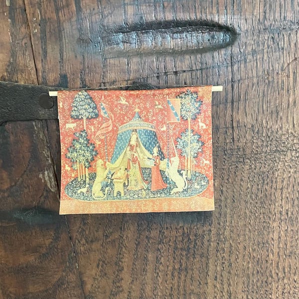 May include: A rectangular wall hanging featuring a printed reproduction of The Lady and the Unicorn tapestry. The scene depicts a central figure in a blue tent, flanked by a lion and a unicorn, surrounded by trees and small animals on a red background.