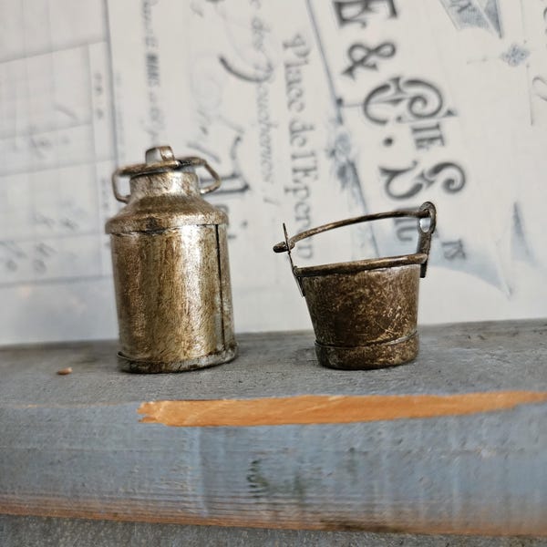 May include: A miniature metal milk can and a small metal bucket with a handle. Both items feature a weathered, distressed gold and brown finish with visible texture, standing side by side as individual decorative pieces for display.