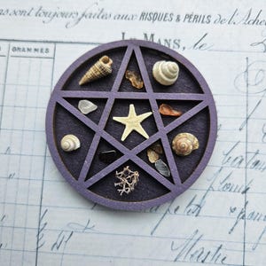 May include: A purple wooden pentagram filled with various seashells, small stones, and a starfish. The pentagram is set against a background of vintage paper with handwritten script. The arrangement includes a variety of natural elements.