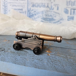 Pode incluir: Um canhão de bronze em miniatura com uma base marrom escura e rodas pretas. O cano do canhão é de uma cor bronze mais clara. O fundo inclui uma superfície de madeira azul e tecido de renda branco.