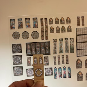 Può includere: Un modello in legno di una torre di chiesa con vetrate colorate. La torre è decorata con un motivo a stella e ha un piccolo campanile in cima. La torre è circondata da un foglio di carta con immagini di vetrate colorate.