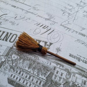 May include: A small broom with a brown wooden handle and straw bristles, secured with black twine. It rests on a vintage paper background featuring faded text and architectural drawings.