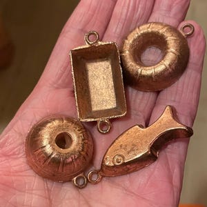 Puede incluir: Cuatro peque&ntilde;os dijes de color cobre en varias formas: una caja rectangular, una rosquilla, una forma redondeada con un agujero y un pez. Cada dije tiene un lazo para sujetarlo a joyas u otras manualidades. Los dijes tienen un acabado met&aacute;lico.