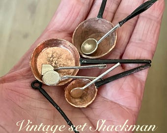 Puede incluir: Un juego vintage de ollas y cucharas de cobre en miniatura con mangos negros. El juego incluye tres ollas pequeñas y tres cucharas. El texto "Vintage set Shackman" es visible en la imagen.