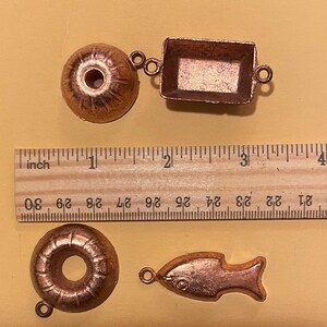 Puede incluir: Cuatro peque&ntilde;os dijes de color cobre: una rosquilla, un pez, un objeto redondo y una bandeja rectangular. Una regla de madera con medidas en pulgadas est&aacute; en el fondo. El dije de rosquilla tiene un agujero en el centro. El dije de pez tiene un detalle de ojo.