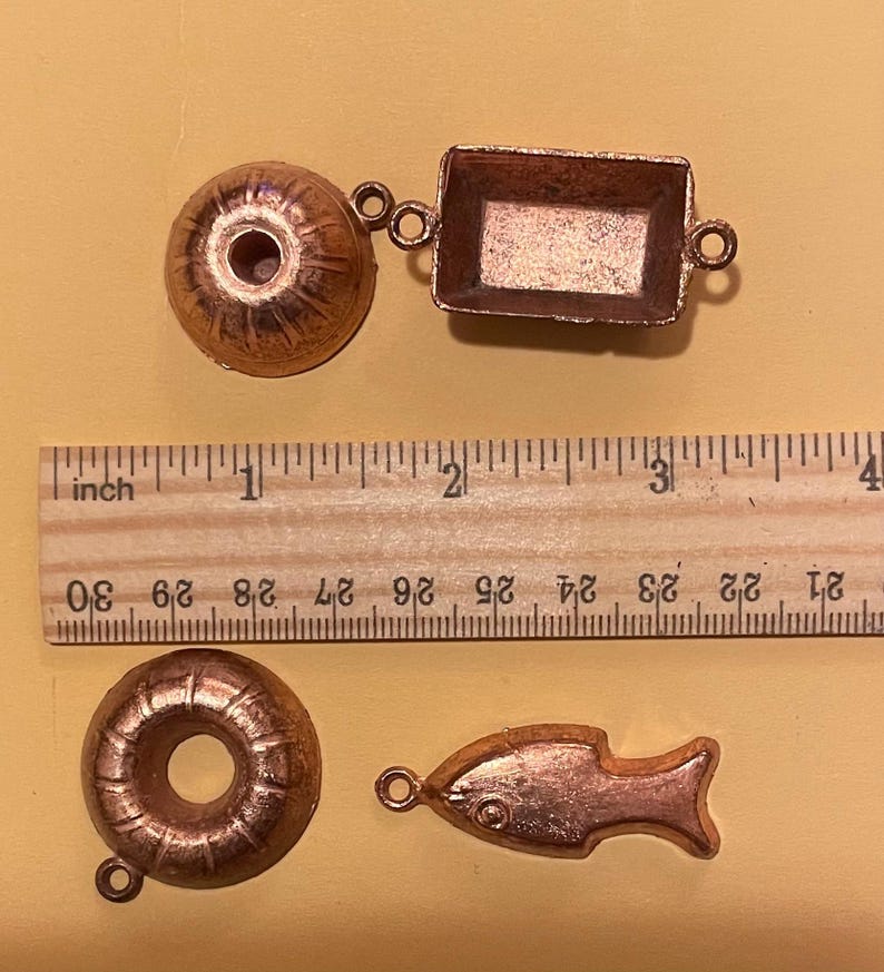 Puede incluir: Cuatro peque&ntilde;os dijes de color cobre: una rosquilla, un pez, un objeto redondo y una bandeja rectangular. Una regla de madera con medidas en pulgadas est&aacute; en el fondo. El dije de rosquilla tiene un agujero en el centro. El dije de pez tiene un detalle de ojo.