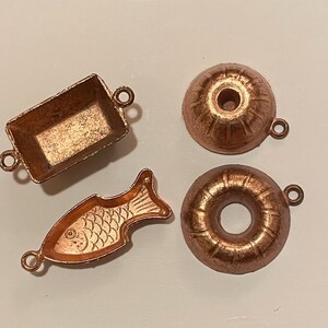 Puede incluir: Cuatro peque&ntilde;os dijes de color cobre en varias formas: una bandeja rectangular, un pez, un bizcocho y una rosquilla. Cada dije tiene un peque&ntilde;o lazo para sujetarlo a joyas u otras manualidades. Los dijes est&aacute;n sobre una superficie clara.