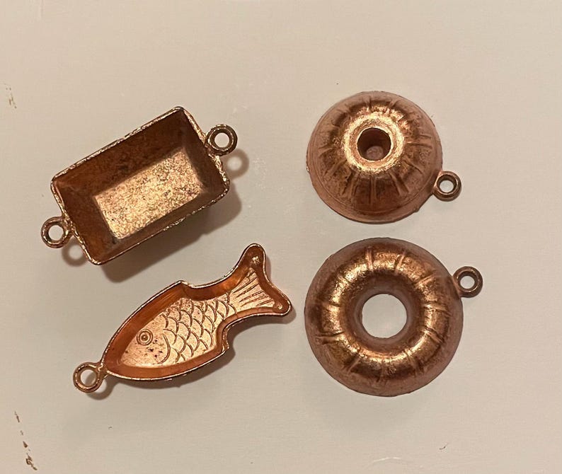 Puede incluir: Cuatro peque&ntilde;os dijes de color cobre en varias formas: una bandeja rectangular, un pez, un bizcocho y una rosquilla. Cada dije tiene un peque&ntilde;o lazo para sujetarlo a joyas u otras manualidades. Los dijes est&aacute;n sobre una superficie clara.