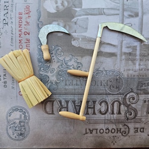 Puede incluir: Una vista cenital presenta herramientas agrícolas en miniatura: una pequeña guadaña, una hoz y un manojo de paja atado con hilo. Las herramientas se colocan sobre un fondo de estilo vintage con texto en francés.