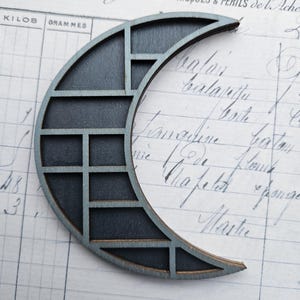 Puede incluir: Recorte de madera en forma de media luna gris oscuro con un diseño geométrico. La luna está sobre un fondo de papel vintage con escritura manuscrita. Mide aproximadamente 10 cm de largo.