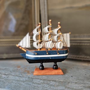 Puede incluir: Maqueta de velero de madera con velas blancas y casco azul y blanco. El modelo tiene tres mástiles y está montado sobre una pequeña base de madera. Un artículo náutico decorativo.