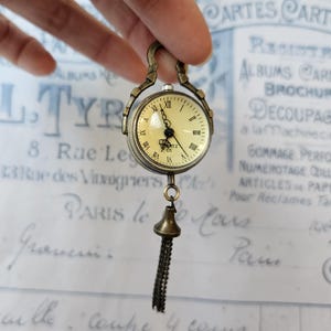 Puede incluir: Un reloj de bolsillo de estilo vintage con esfera crema, manecillas negras y números romanos. El reloj tiene una caja de metal color bronce y una cadena decorativa con un pequeño dije en forma de campana. La palabra "QUARTZ" está impresa en la esfera.