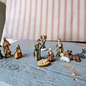 Puede incluir: Un belén en miniatura con figuras pintadas. La escena incluye a los Reyes Magos, María, José, el niño Jesús, un ángel, un pastor, un cordero, un burro y una vaca. Las figuras están colocadas sobre una superficie de madera azul.