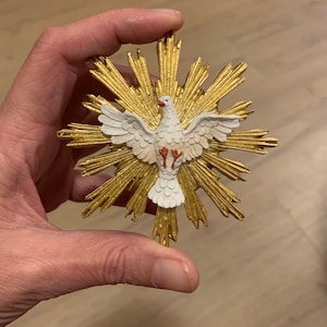 Op de afbeelding: Een witte duif met rode poten, met gespreide vleugels, bevindt zich in het midden van een gouden zonneschijn. De duif is een symbool van de Heilige Geest.