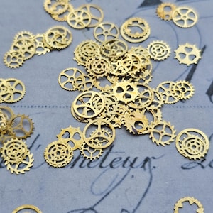 Puede incluir: Una colección de pequeños engranajes metálicos dorados dispersos sobre una superficie azul claro con escritura descolorida. Los engranajes varían en tamaño y diseño, con detalles intrincados y una estética vintage. Ideal para manualidades o proyectos steampunk.