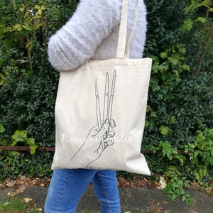 K&ouml;nnte beinhalten: Eine wei&szlig;e Canvas-Tragetasche mit einer schwarzen Linienzeichnung einer Hand, die drei Pinsel h&auml;lt. Der Text "Rosanne Watts" ist auf die Tasche gedruckt.