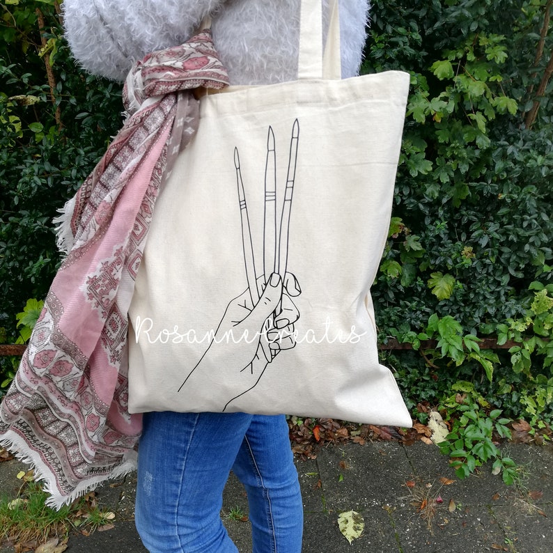 K&ouml;nnte beinhalten: Eine wei&szlig;e Canvas-Tragetasche mit einer schwarzen Linienzeichnung einer Hand, die Pinsel h&auml;lt. Der Text "Rosanne Creates" ist auf die Tasche gedruckt.