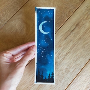 Puede incluir: Una pintura de acuarela de una luna creciente en un cielo nocturno azul oscuro con estrellas blancas. La luna es blanca y el cielo es azul oscuro con un degradado de azul más claro en la parte inferior. Hay árboles verde oscuro en la parte inferior de la pintura.
