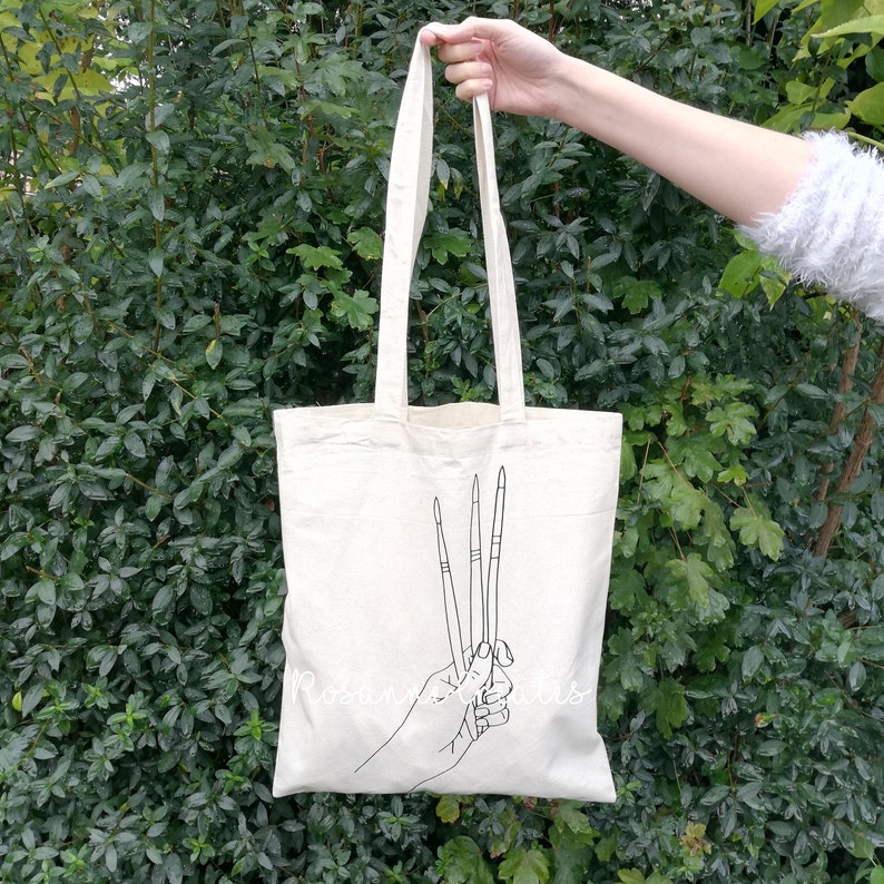K&ouml;nnte beinhalten: Eine wei&szlig;e Stofftasche mit einer schwarzen Linienzeichnung einer Hand, die drei Pinsel h&auml;lt.