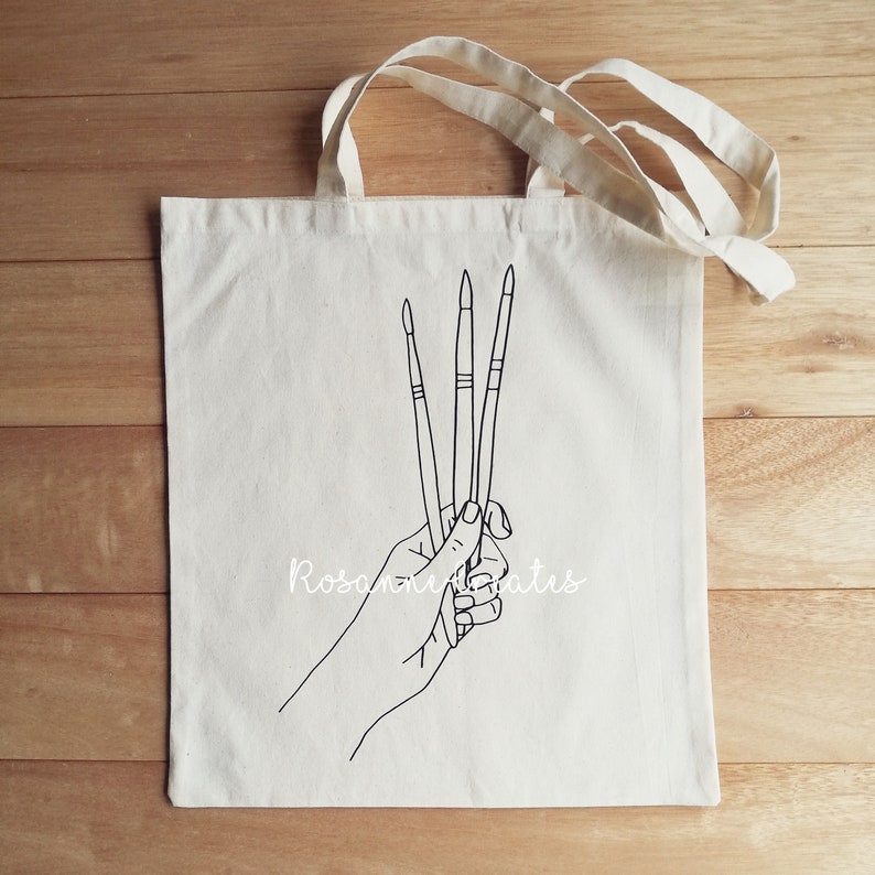 K&ouml;nnte beinhalten: Eine wei&szlig;e Canvas-Tragetasche mit schwarzer Linienzeichnung einer Hand, die drei Pinsel h&auml;lt. Der Text "Rosanne Creates" ist auf die Tasche gedruckt.