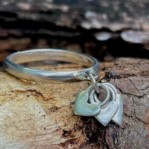 May include: A silver ring with three heart-shaped charms hanging from it. The ring and charms are made of a shiny metal and rest on a textured, brown surface. The hearts are connected by small rings.