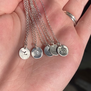 May include: A set of six silver necklaces with different charms. The charms are small, round, and feature various designs, including a sun, a moon, a bird, a star, and a flower.