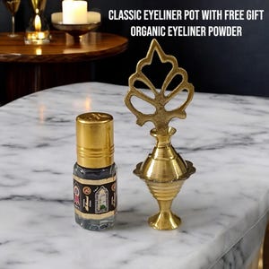 May include: A gold metal eyeliner pot with a decorative top and a small glass bottle of black eyeliner. The bottle has a gold cap and a label with Arabic text. The text "CLASSIC EYELINER POT WITH FREE GIFT ORGANIC EYELINER POWDER" is visible above the items.