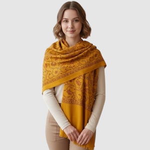 Op de afbeelding: Een mosterdgele sjaal met een donkerbruin bloemen- en rankenpatroon. De sjaal is gedrapeerd over de schouders en borst, met een brede rand voorzien van gedetailleerde botanische motieven en een zachte, gladde textuur over de gehele stof.