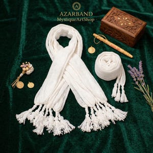 Puede incluir: Bufanda blanca con borlas, exhibida sobre una superficie de terciopelo verde. La bufanda está drapeada y enrollada, mostrando su textura y diseño. Elementos adicionales incluyen una caja de madera, un bolígrafo y elementos decorativos.