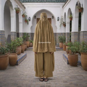Può includere: Un burqa color beige e un completo pantalone abbinato. Il burqa presenta una copertura per il viso ricamata e un design a mantello fluente. L'outfit è indossato con sandali neri. Lo sfondo include un cortile con piante in vaso e dettagli architettonici.