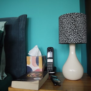 Op de afbeelding: Een nachtkastje met een witte lamp met een zwart-witte lampenkap, een zwarte telefoon, een doos tissues, een boek met de titel "The Hunger Games" en een bril.